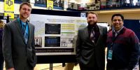 Two students and professor posing by their presentation at the National Conference of Undergraduate Research