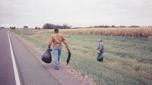 Two people with garbage bags picking up trash on the side of the road near a field