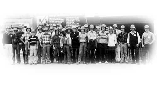 A black and white photo of people working in construction during the 1970s posing outside of the construction site