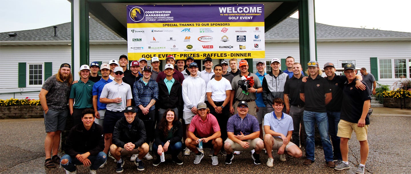 Students gathered together for a picture underneath the Construction Management Golf event poster at the Construction Management Golf event