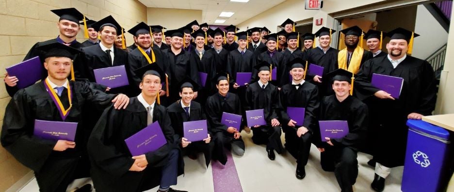 Spring 2019 Construction Management graduates posing in the hallway wearing black graduation gowns and caps with purple graduation folder in their hands