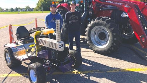 Two Maverick One students posing outside in a parking lot next to a red tractor and their designed competitive tractor